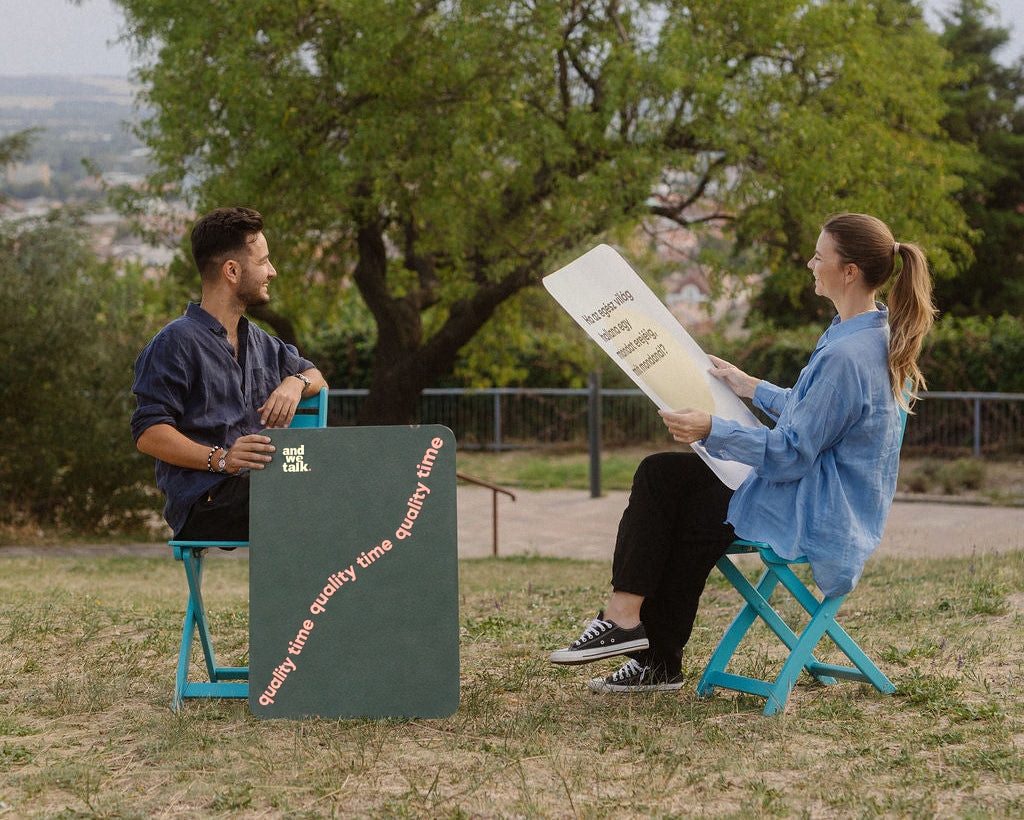 Two people talking under the trees in a park, holding large Quality Time cards